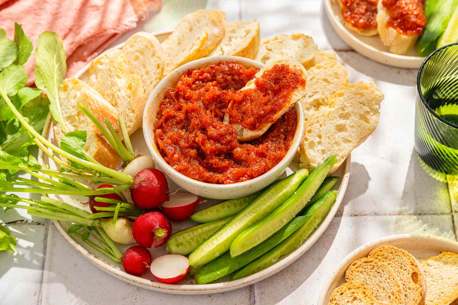 Side view of honey chorizo spread in a bowl surrounded by radishes, cucumber spears and sliced baguette, with one baguette slice topped with the spread