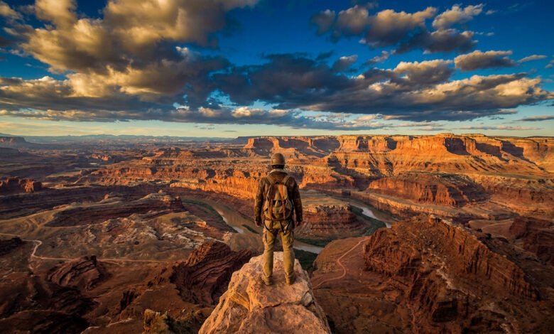 Un aventurier équipé d'un sac à dos se tient debout sur un pic rocheux dominant un vaste canyon au coucher du soleil.