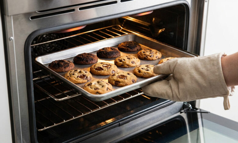 Une main gantée retire une plaque de cookies d'un four en acier inoxydable. Les cookies présentent des niveaux de cuisson différents, du doré au trop cuit.